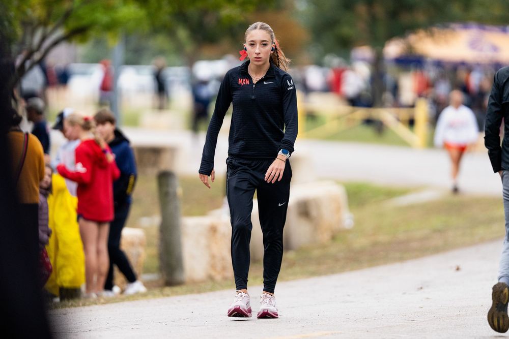 Female cross country runner in black Nike gear walking on a path at an outdoor athletic event.