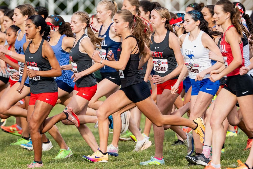 Female cross country runners bursting from the starting line at a competitive high school race.