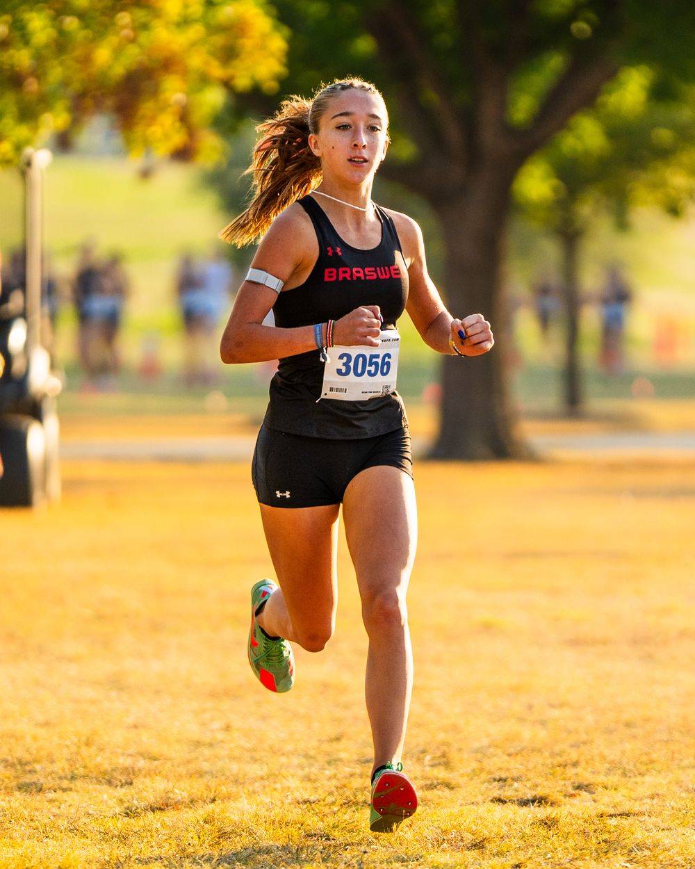 Female Braswell cross country runner wearing bib 3056 racing through a sunlit park during competition.