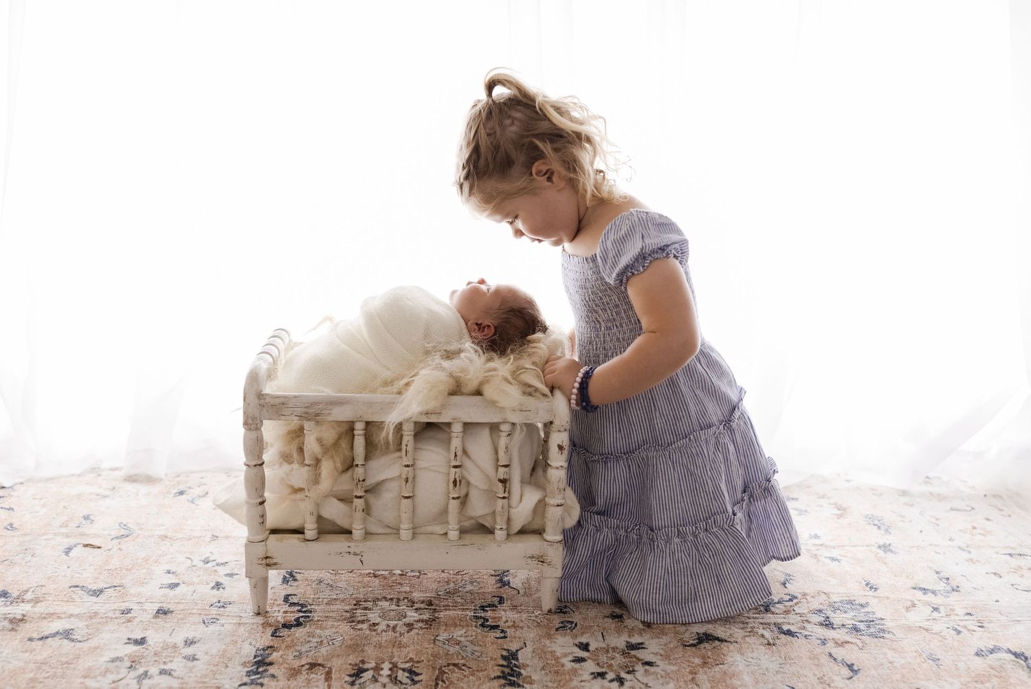 Proud big sister in a nautical dress peers over her newborn sister's crib, captured by Charlottesville baby and family photographer.