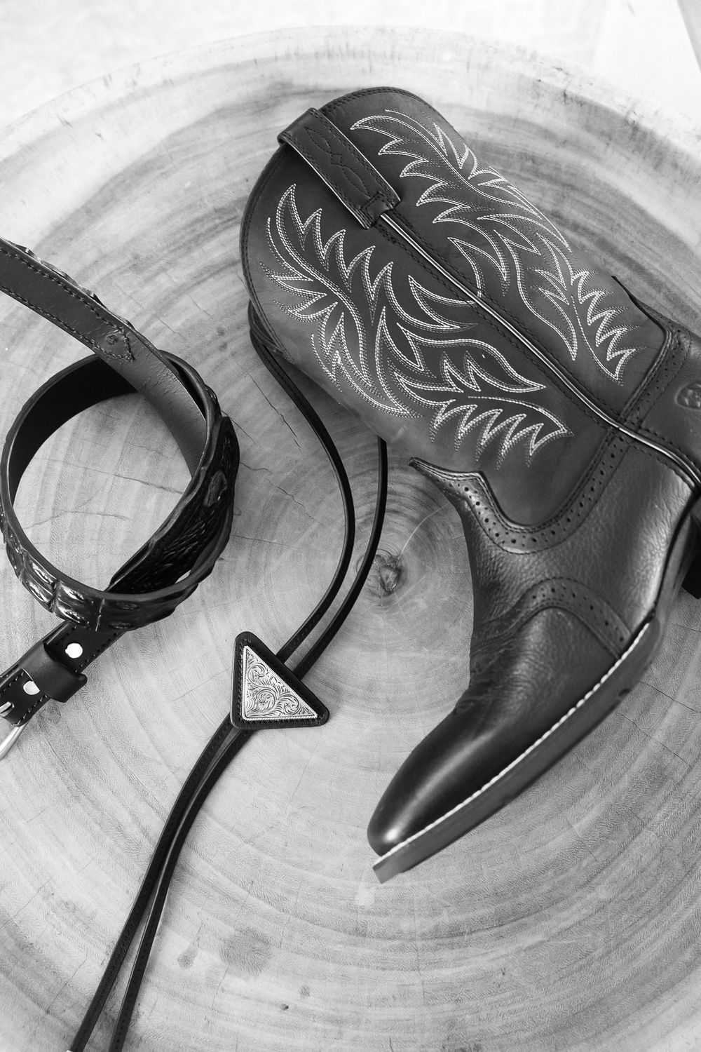 Black and white photo of a cowboy boot with embroidered details beside a leather belt and bolo tie on a wood stump.
