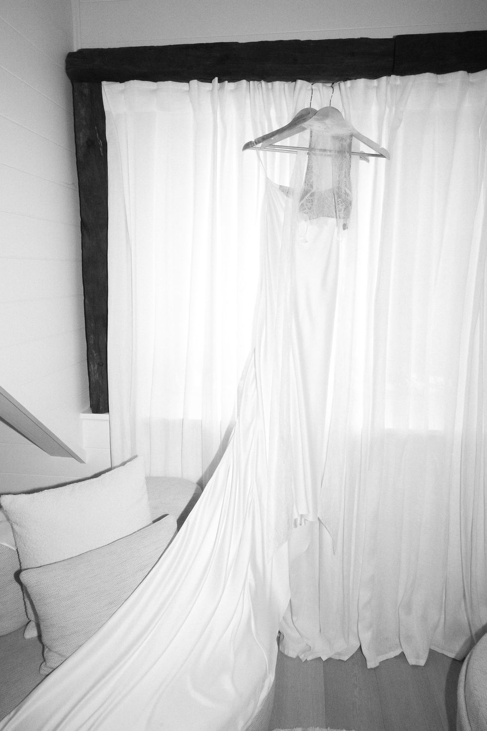 Elegant white wedding dress hanging on a wooden beam by a window with sheer curtains in a black and white photo.