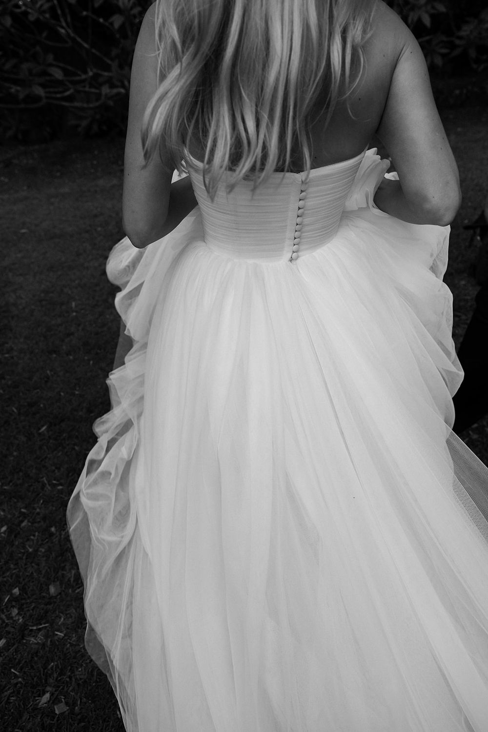 Black and white photo of a bride in a strapless ball gown wedding dress with tulle skirt, viewed from behind.