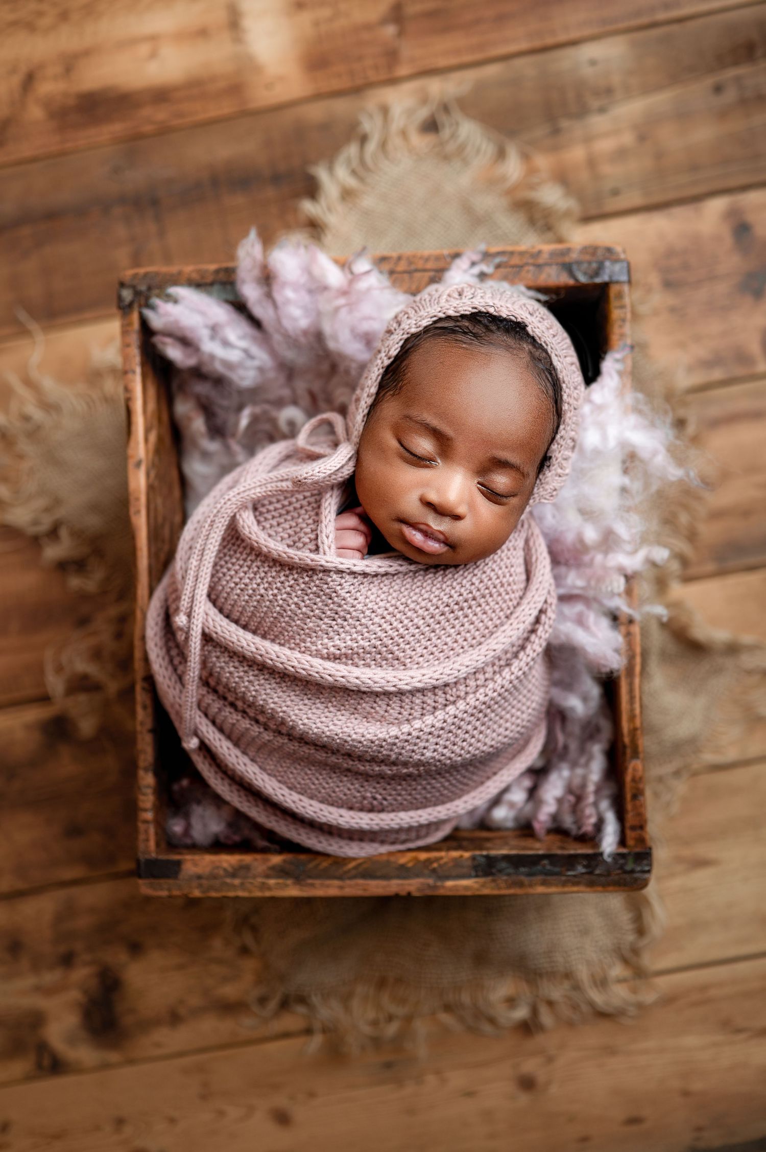 Sleeping newborn baby wrapped in teal swaddle inside a wooden heart-shaped bowl on rustic wood floor.