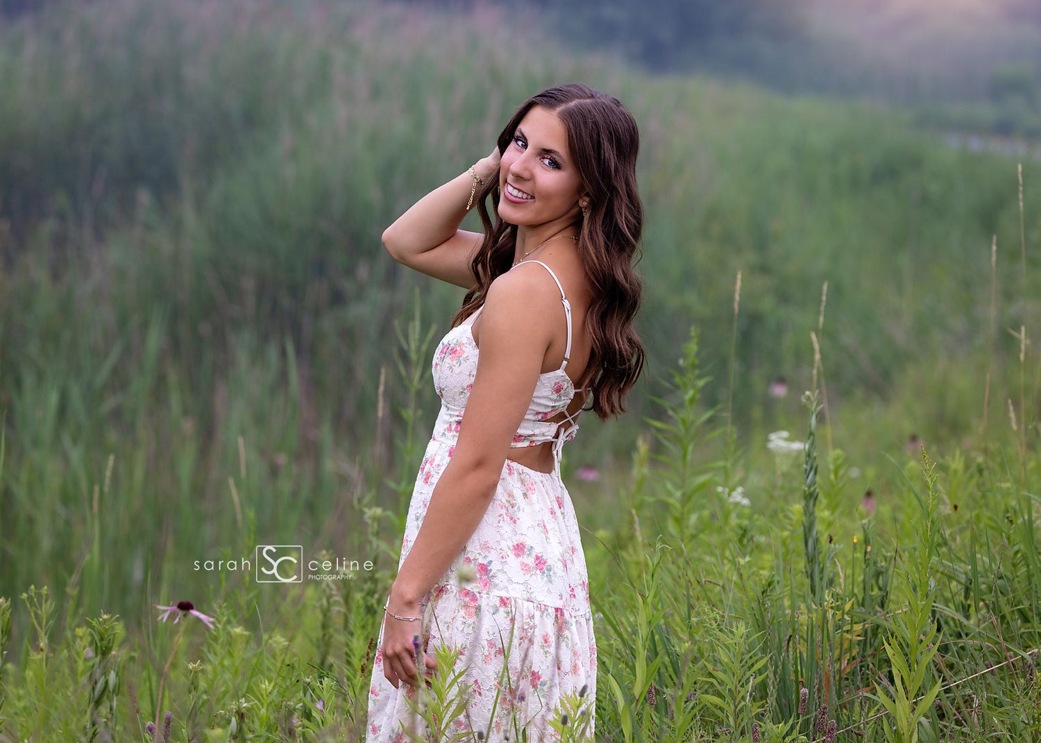 Young woman in floral sundress smiling in lush green field, long brunette hair, outdoor senior portrait photography.