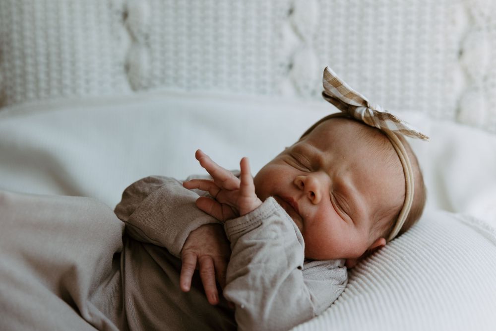 Peaceful newborn baby sleeping in white bedding, wearing beige outfit and bow headband, with tiny hand raised.