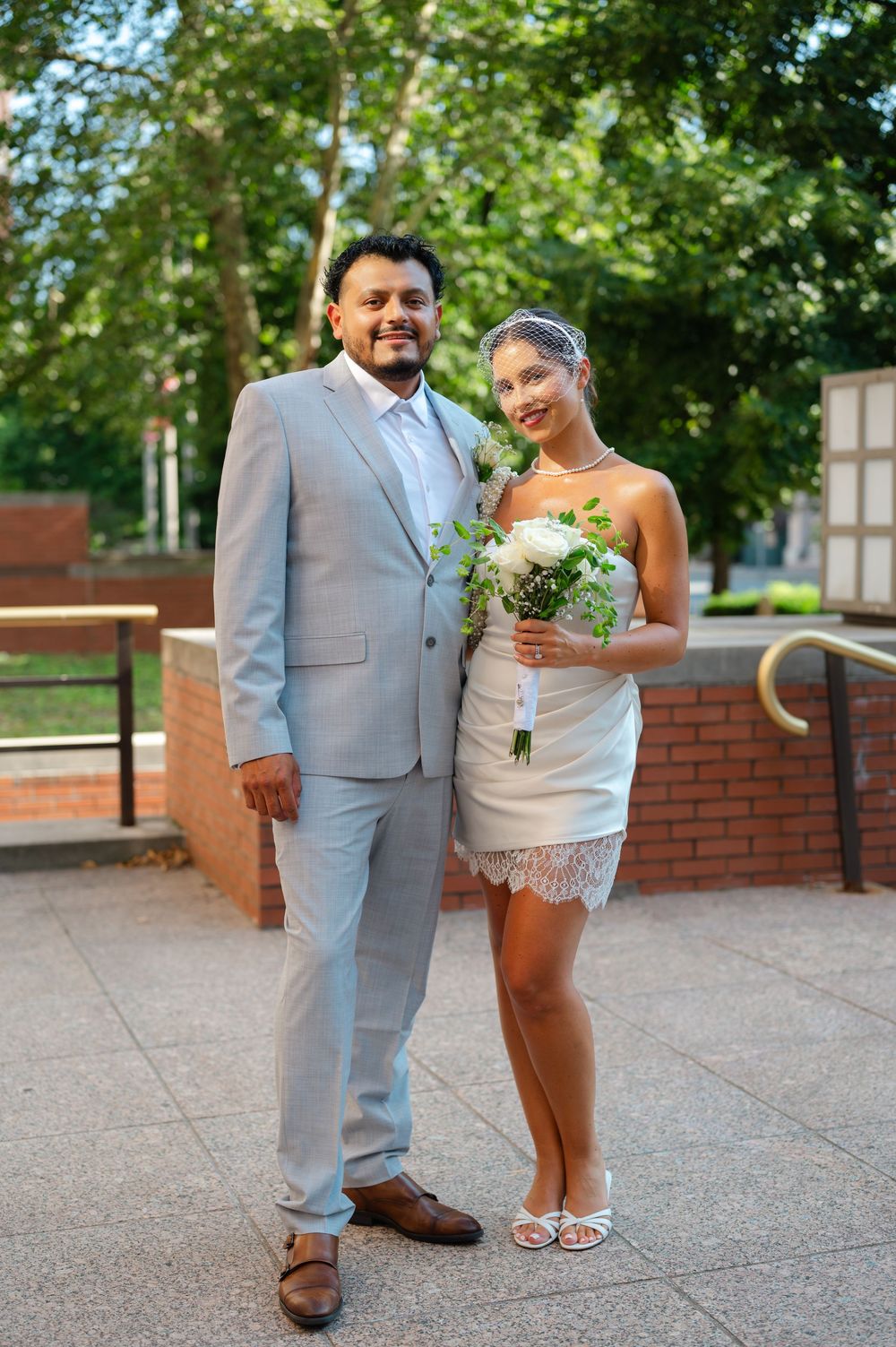 Newlywed couple posing outdoors, groom in gray suit and bride in short white lace dress holding bouquet.
