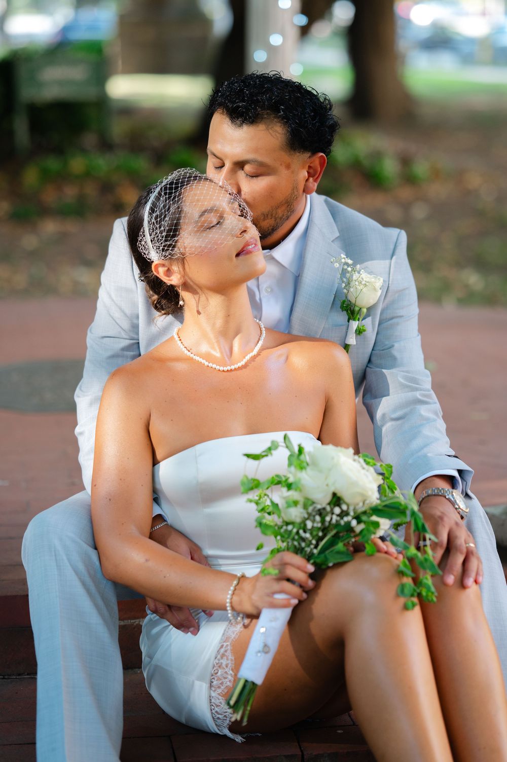 Bride and groom share a tender kiss while seated outdoors, bride holding white bouquet wearing birdcage veil and pearl necklace.