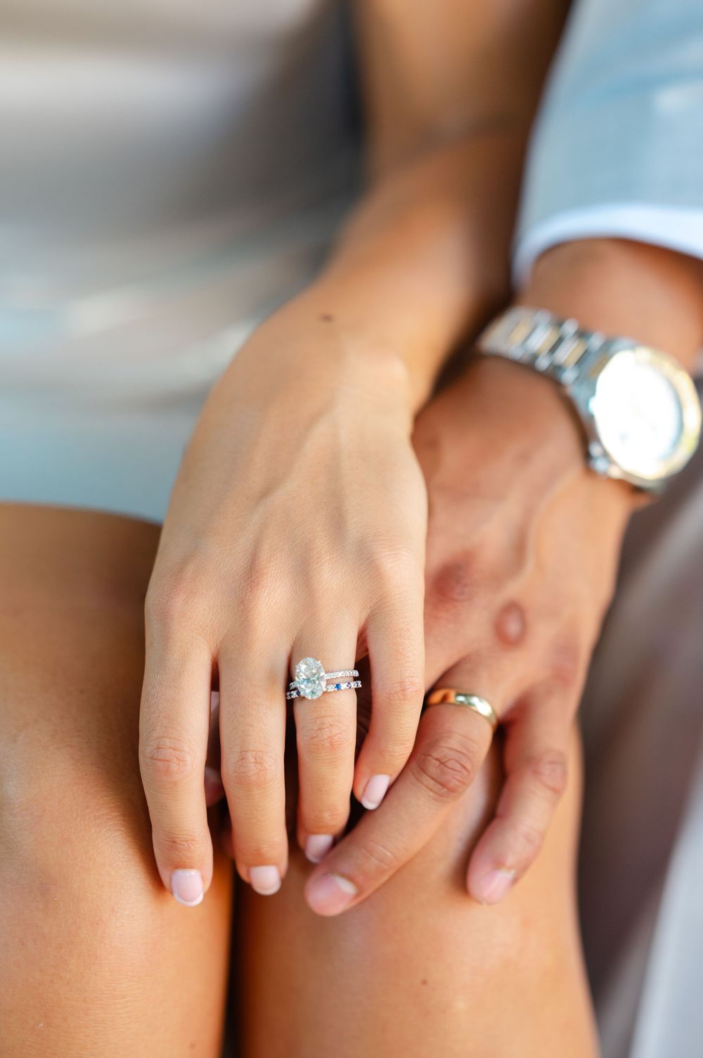 Close-up of a woman's hand wearing a diamond engagement ring and wedding band, with a man's hand wearing a watch beneath.