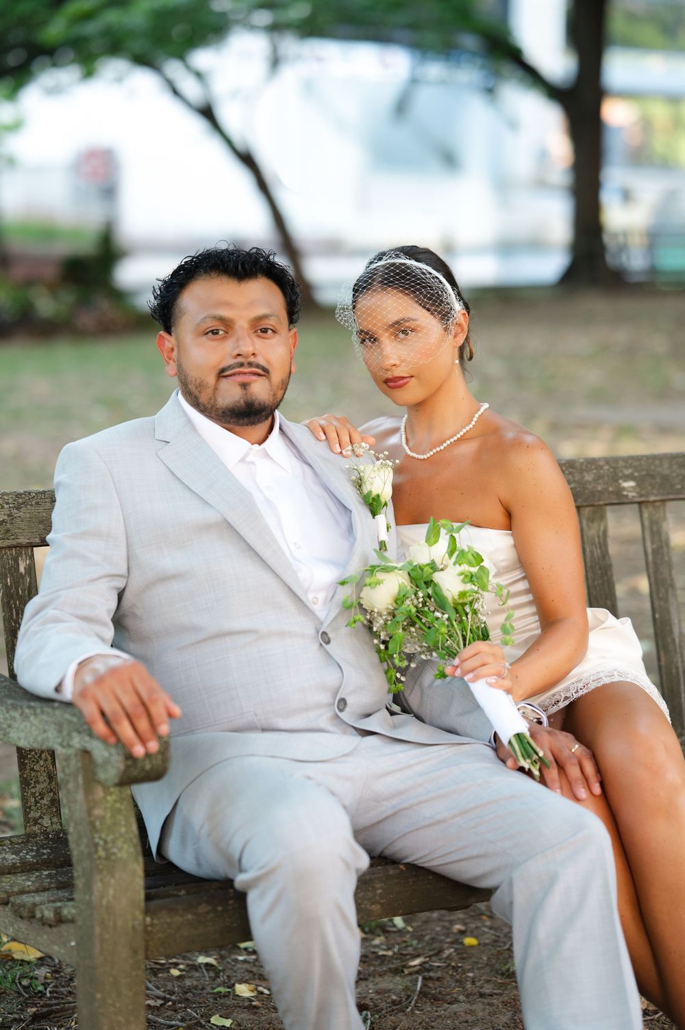 Newlywed couple sitting on a park bench, groom in gray suit, bride in white dress holding a bouquet.