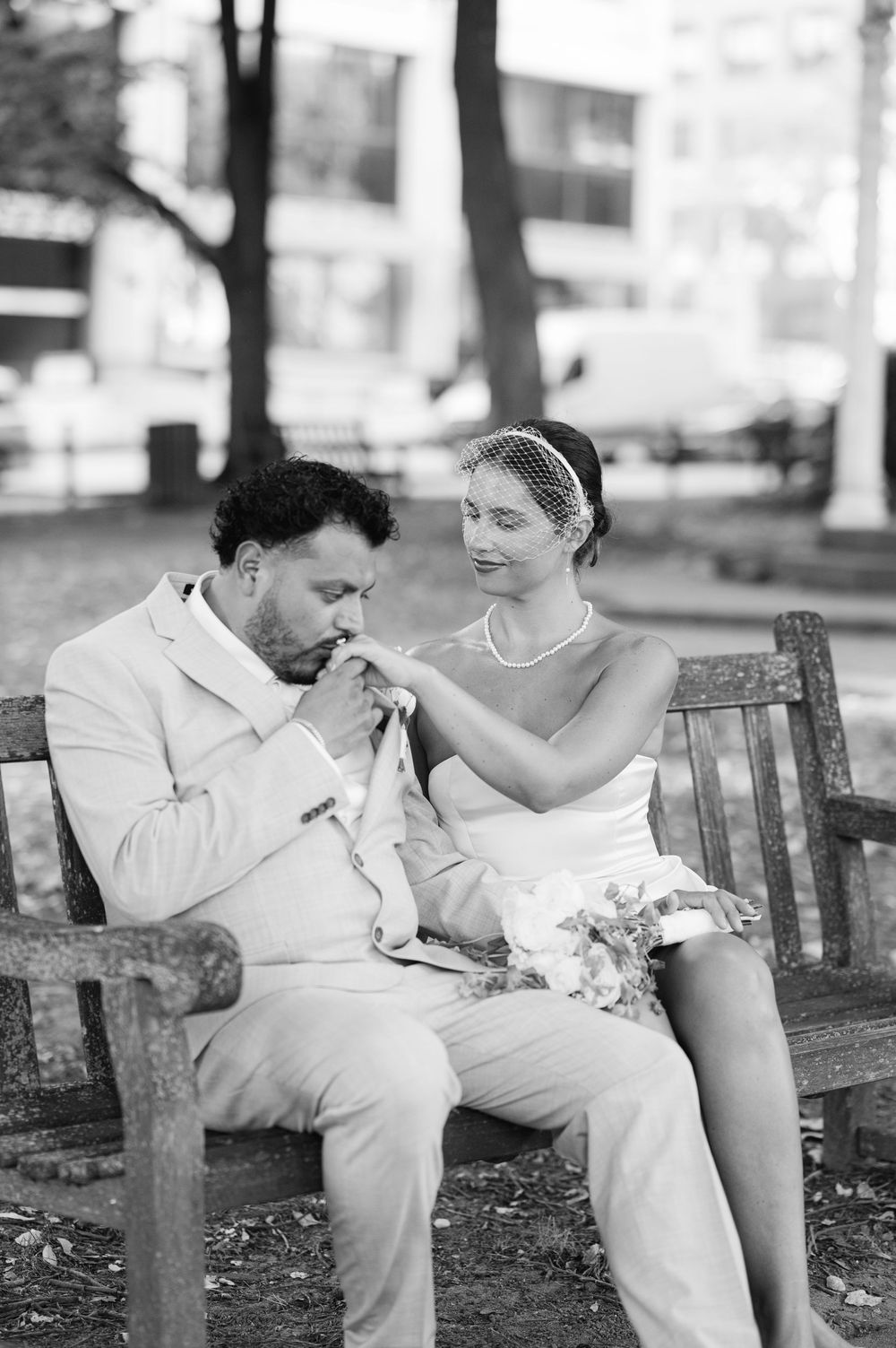 Black and white photo of a groom kissing bride's hand on a park bench, both in elegant wedding attire.