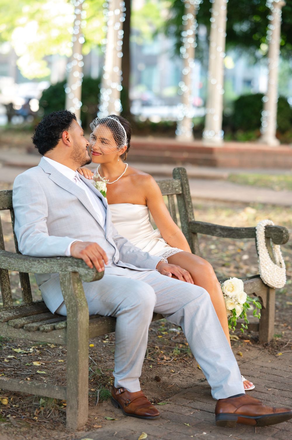 A bride and groom in elegant wedding attire sit closely on a park bench, gazing at each other with string lights behind them.
