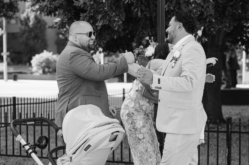 Black and white photo of two men in suits greeting each other at an outdoor wedding ceremony.