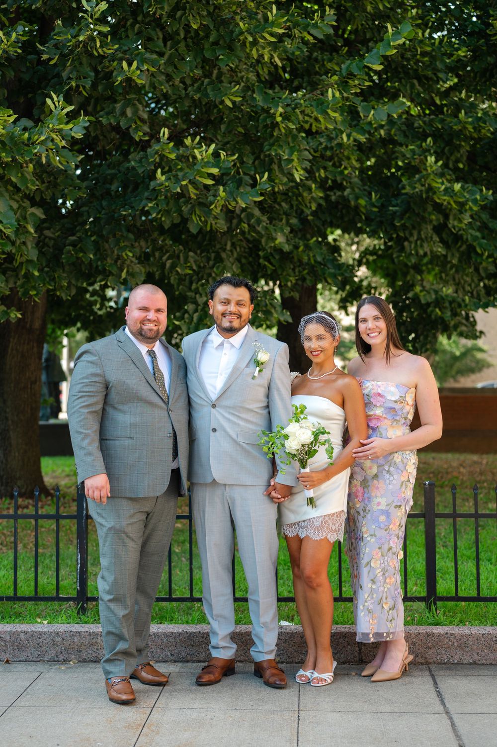 Wedding party of four posing outdoors near iron fence and lush green trees, featuring bride in white lace dress holding bouquet.
