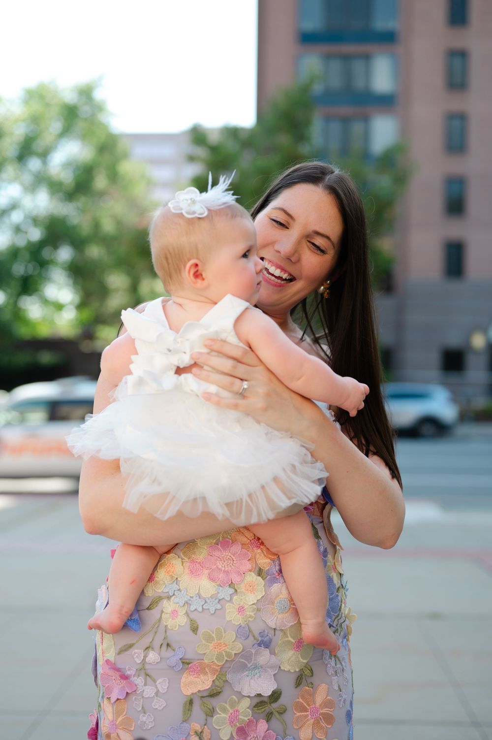 A smiling brunette woman in a floral dress holds a laughing baby girl in a white tutu dress outdoors.