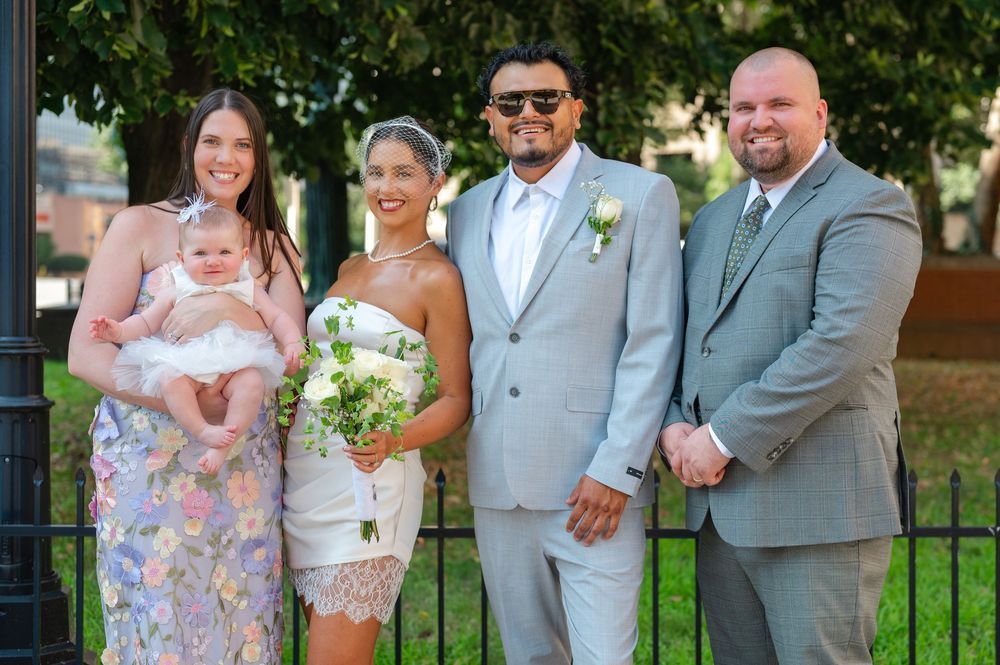 Wedding couple in gray suit and white dress poses with three guests outdoors, bride holding bouquet.