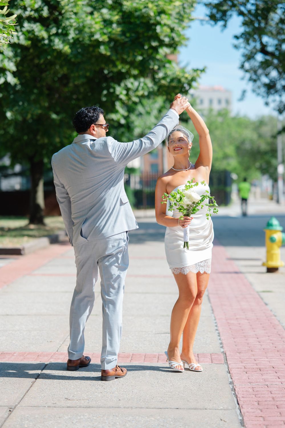 Groom twirling bride in white mini dress on sunny sidewalk during outdoor wedding celebration.