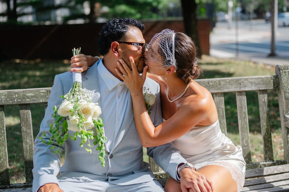 Bride and groom share a kiss on a park bench, bride holding white bouquet, groom in light blue suit.