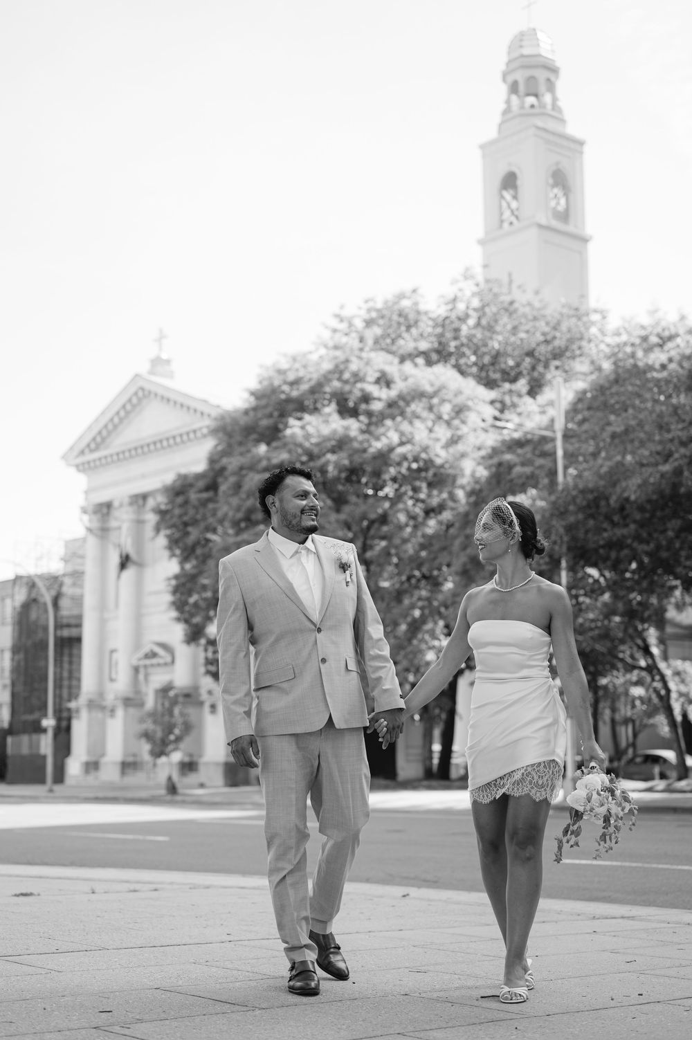 Bride and groom walking hand in hand in black and white photo with historic church in background.