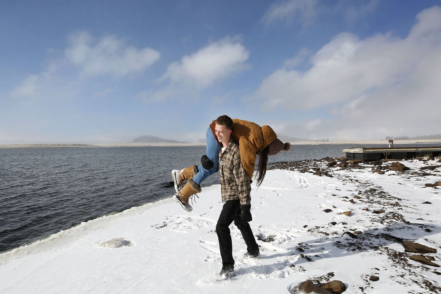 A man in a plaid jacket carries a laughing woman on his back along a snowy lakeshore under a partly cloudy sky.
