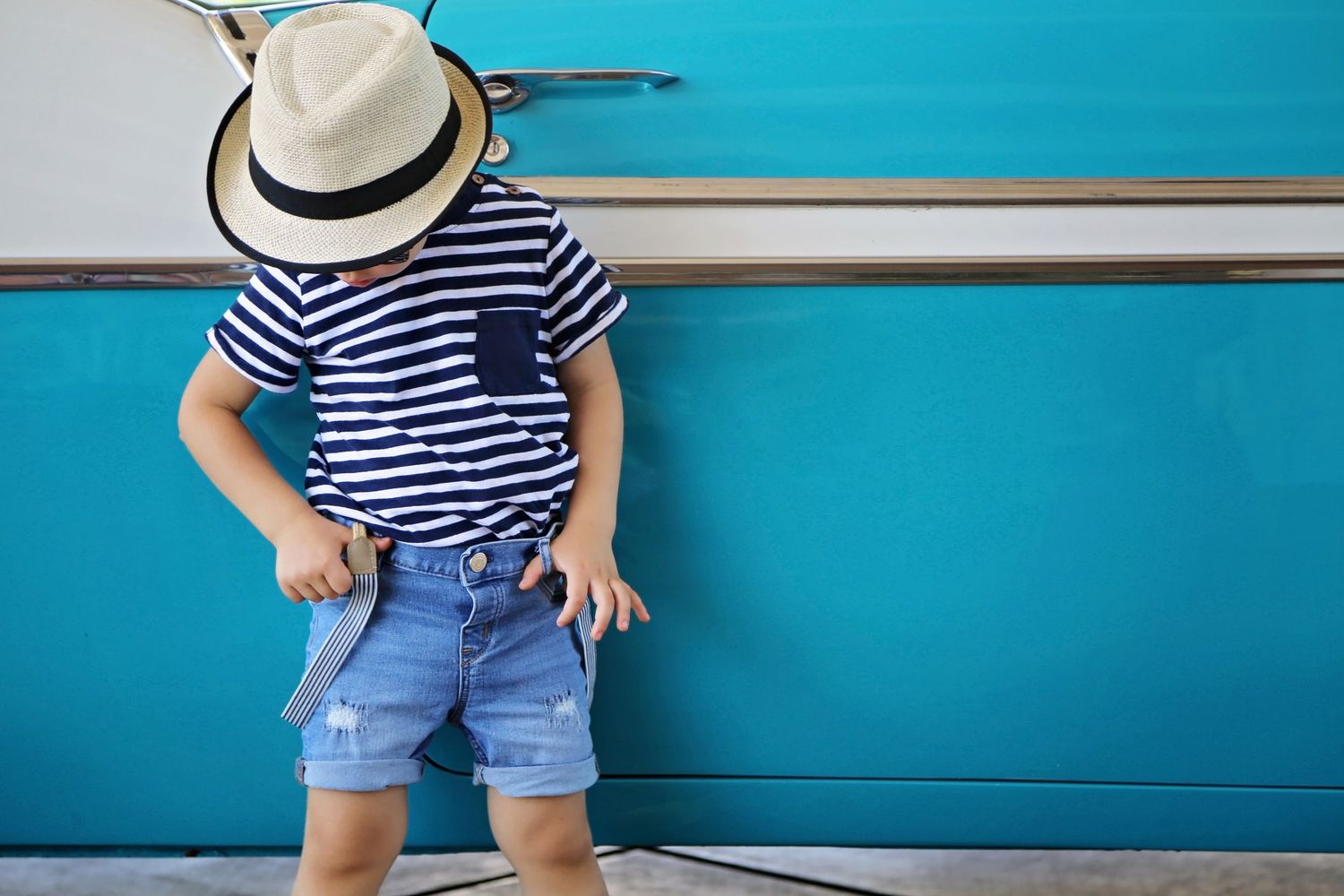 A young boy in a straw hat, striped shirt and denim shorts stands against a turquoise vintage van, adjusting his belt.