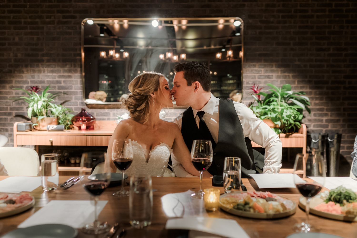 Bride and groom sharing a romantic kiss at a candlelit wedding reception table with red wine glasses.