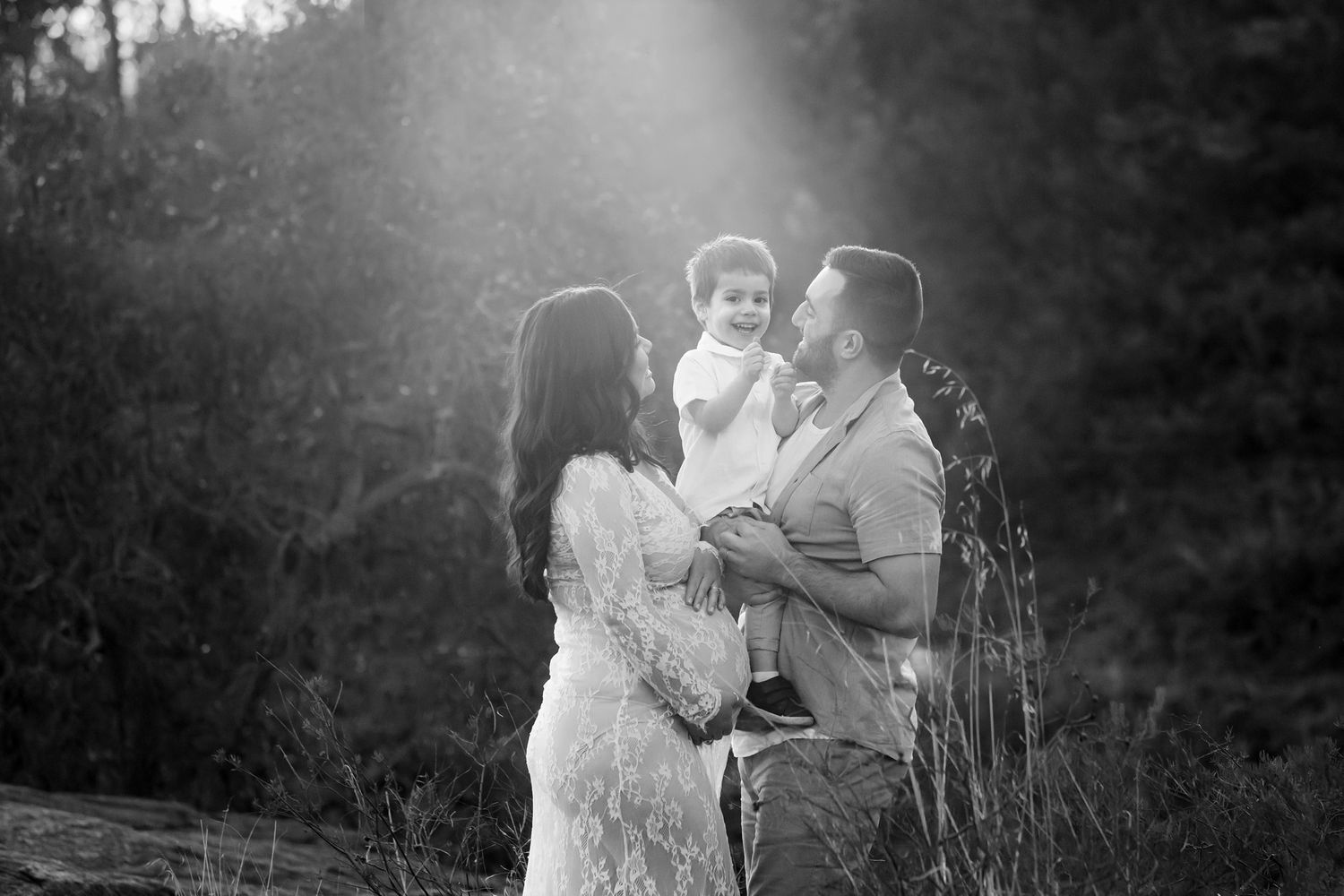 Black and white family portrait of parents holding toddler outdoors with sunlight filtering through trees.