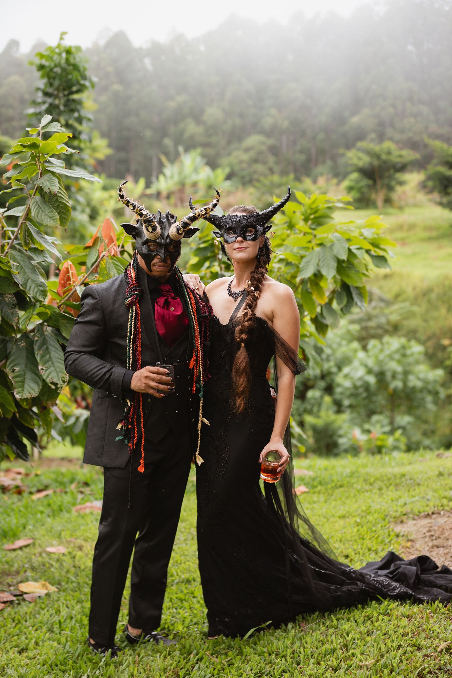 Couple in black outfits and horned masks standing in a lush tropical garden.