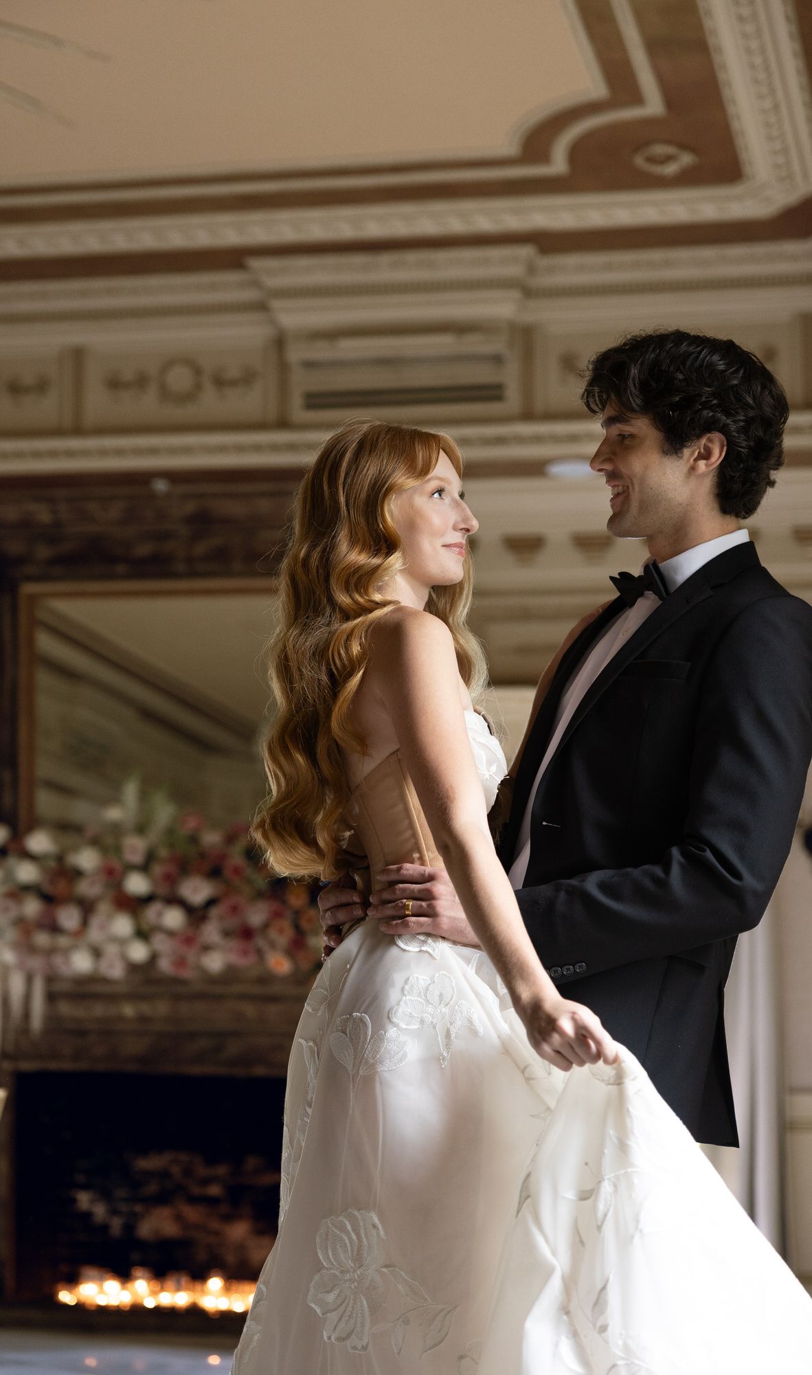 Elegant couple dancing at a wedding, bride in white gown with floral details, groom in black tuxedo, ornate ballroom.