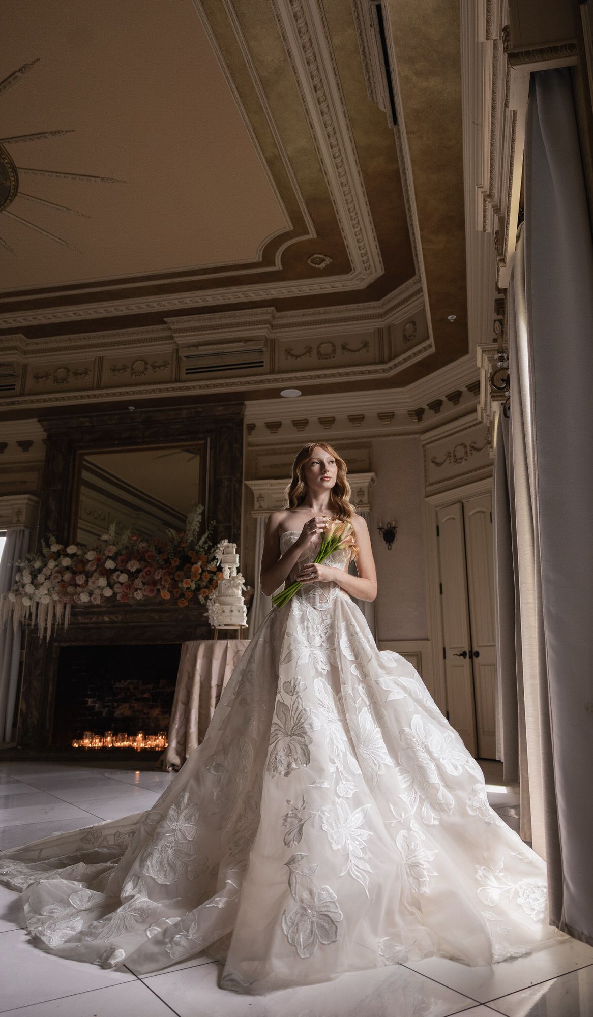 Bride in elegant lace ball gown holding bouquet in ornate room with decorative ceiling and fireplace.