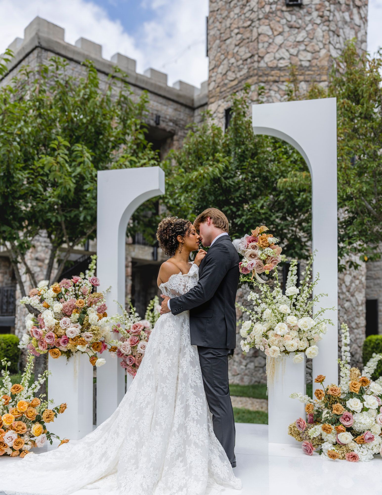 Bride and groom embrace before white arch backdrop with colorful floral arrangements at elegant castle wedding venue.