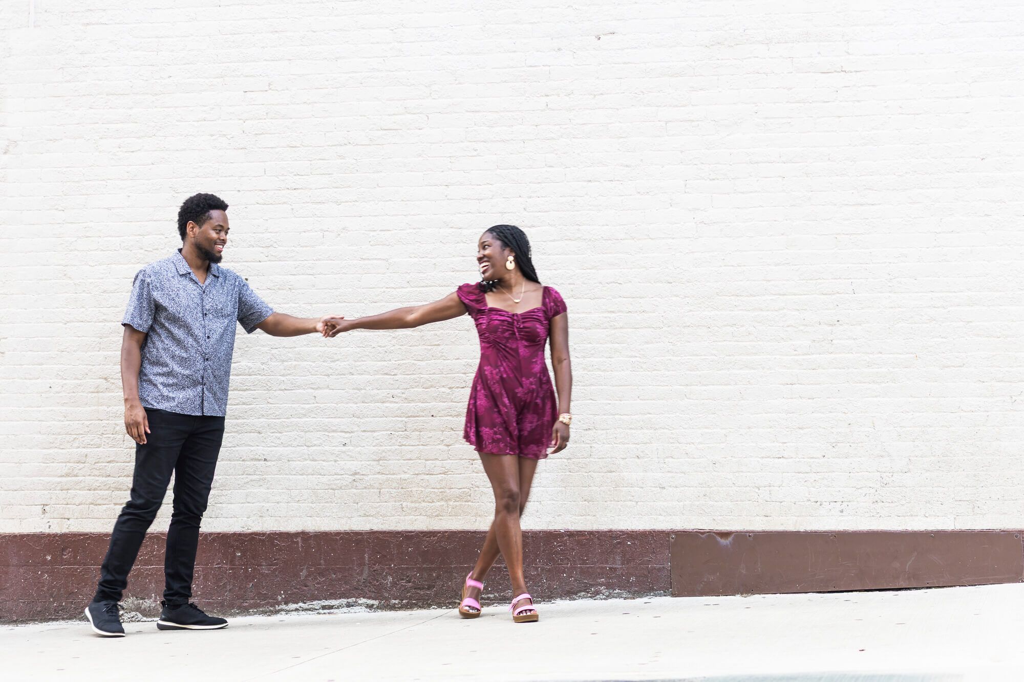 A young Black couple holds hands and smiles at each other against a white brick wall outdoors.