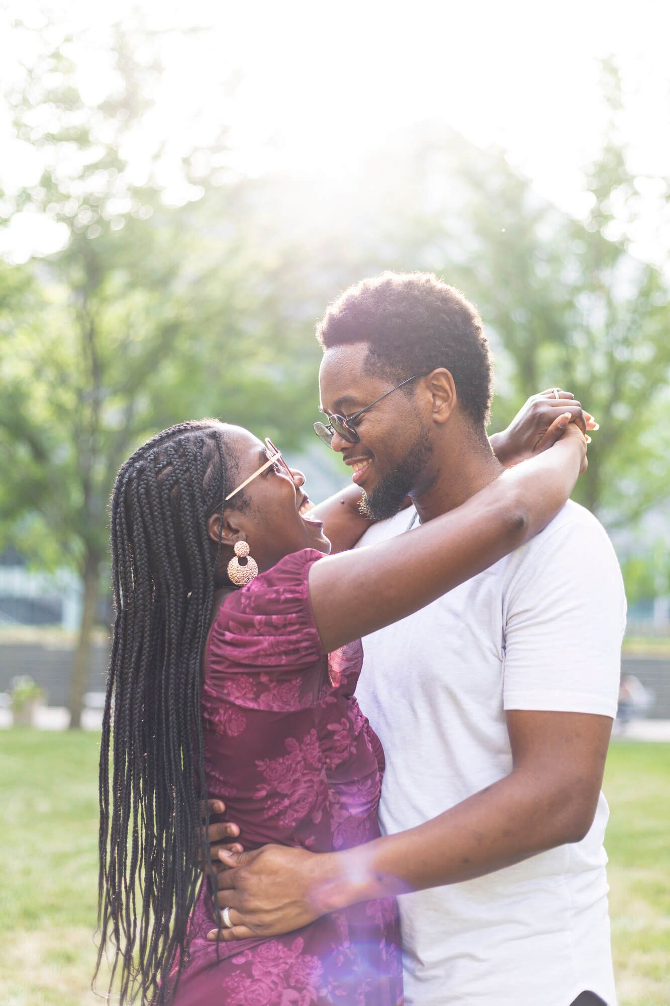 A joyful Black couple embraces and smiles at each other outdoors in a sunlit park, woman in floral dress with braids.