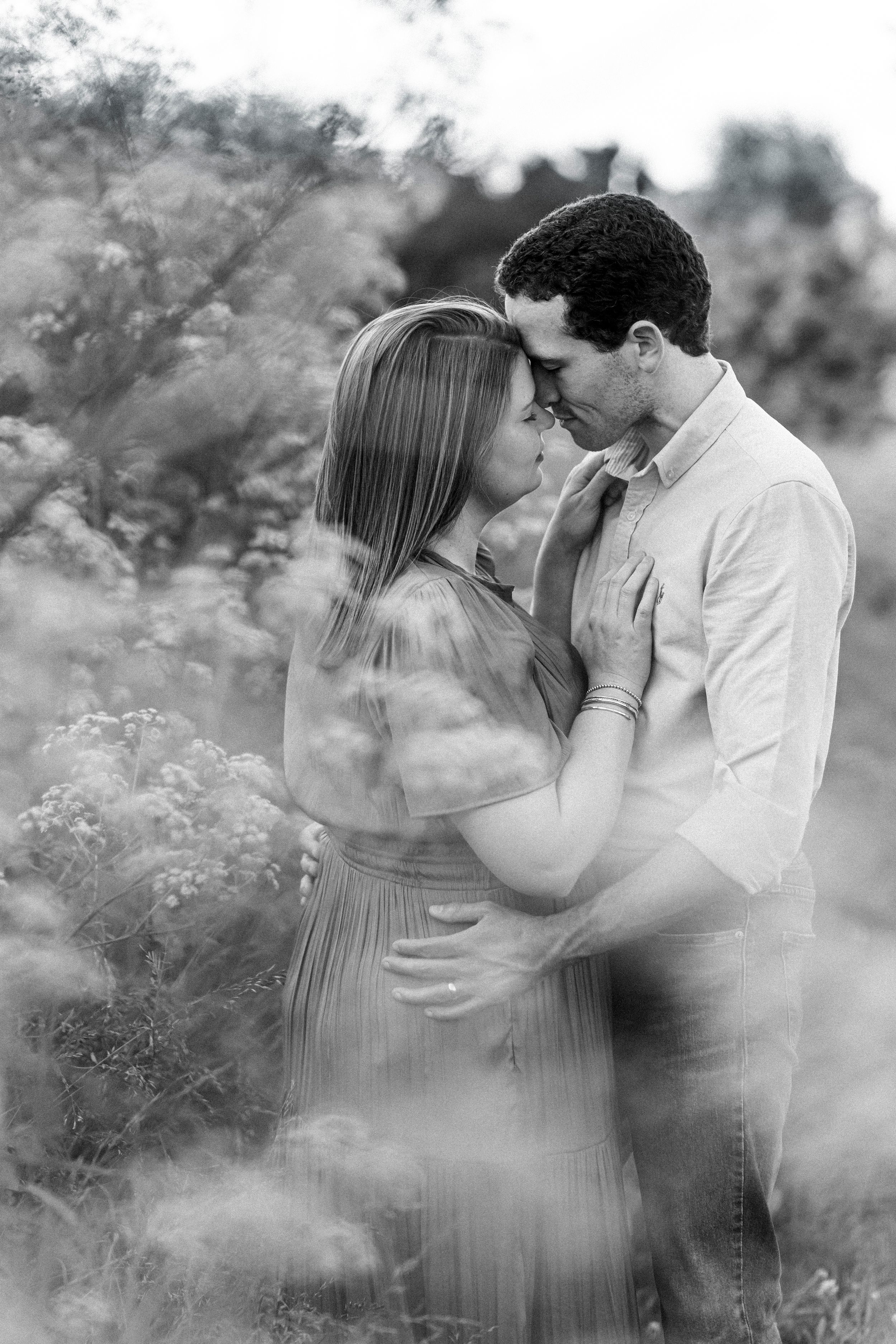 Black and white photo of a couple embracing tenderly in a field of wildflowers, foreheads touching romantically.