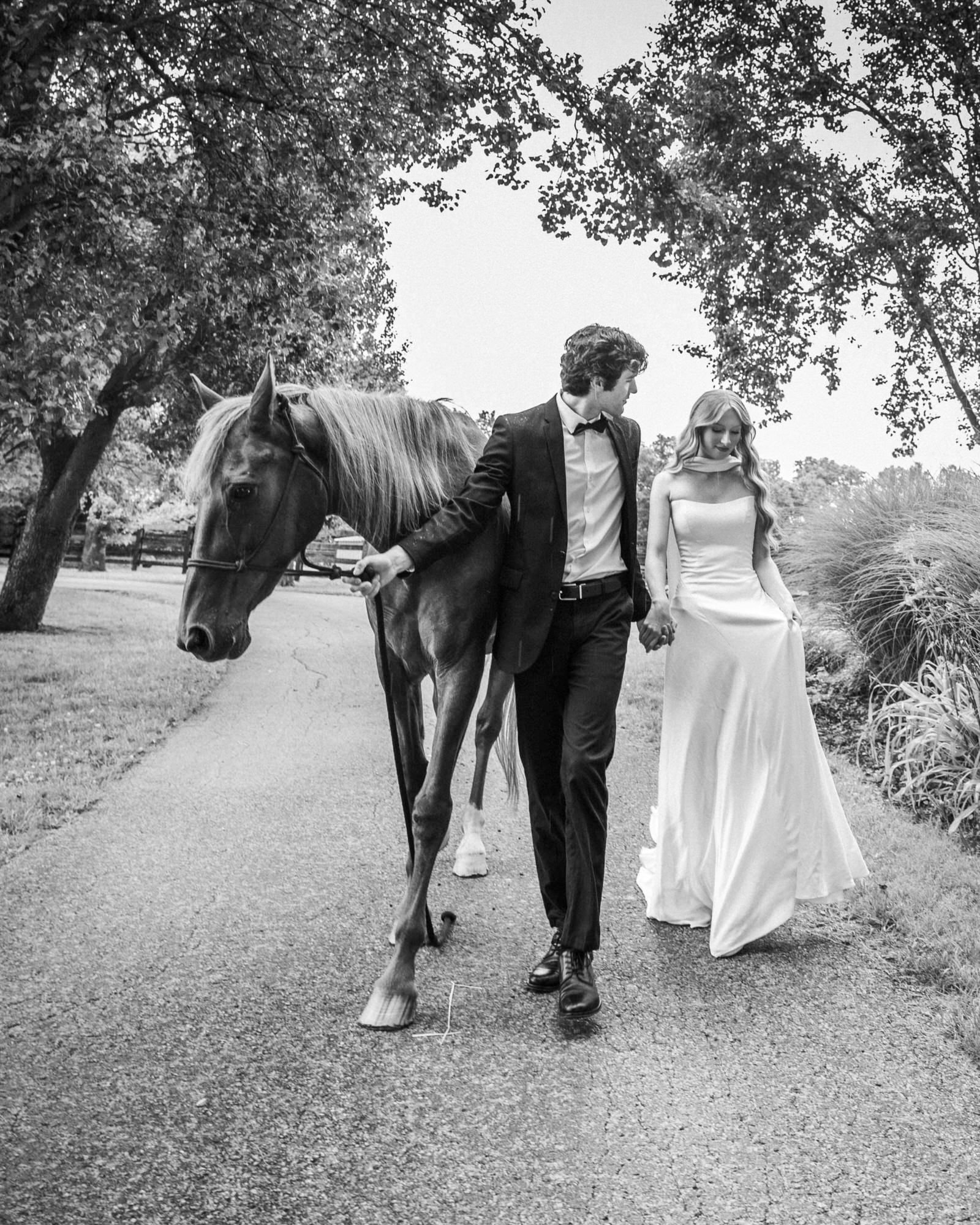 Black and white photo of a bride and groom walking a horse down a tree-lined path on their wedding day.