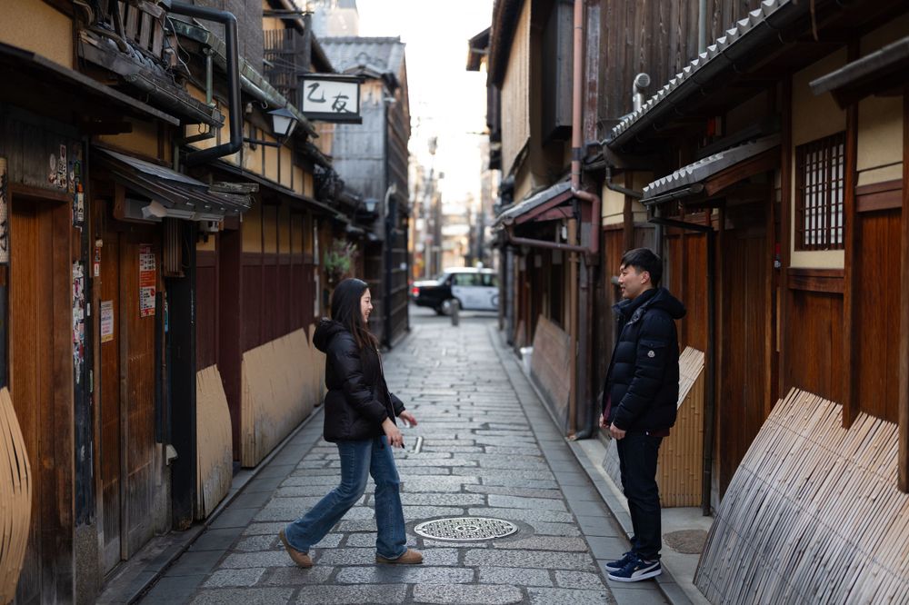Two people face each other on a narrow cobblestone alley lined with traditional Japanese wooden buildings in Kyoto.