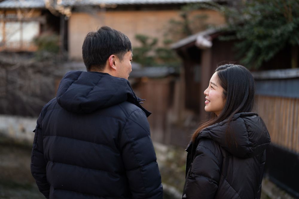Young Asian couple share a warm smile while standing outdoors near traditional Japanese architecture in winter.