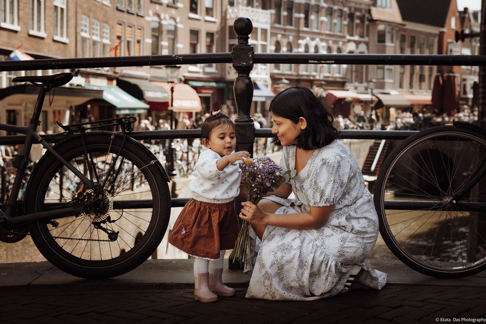 Mother crouching to give purple flowers to toddler daughter on Amsterdam canal bridge with bicycles.