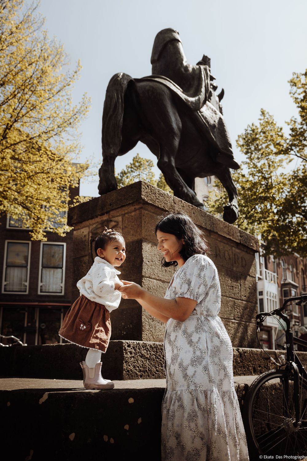 Mother and toddler playing near an equestrian statue in Amsterdam on a sunny day.