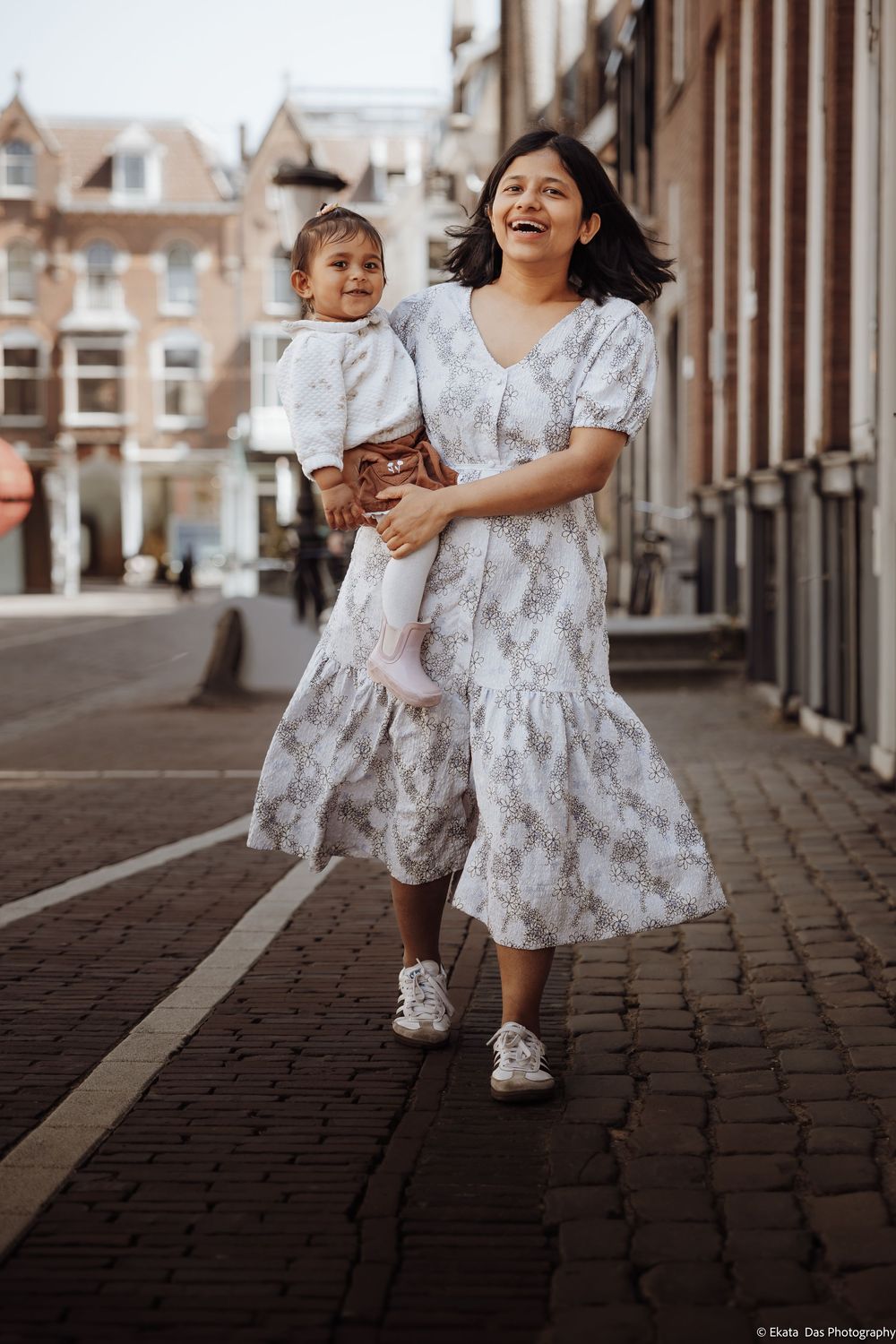 A joyful mother in a floral dress carries her toddler while walking on a cobblestone street in Amsterdam.