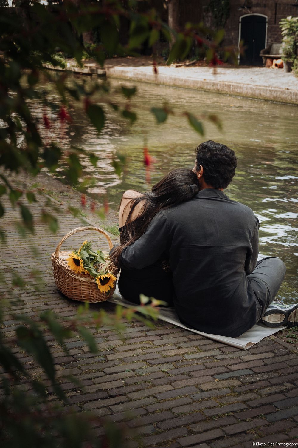 Couple sitting together on brick canal edge with sunflower picnic basket, sharing a romantic outdoor moment.