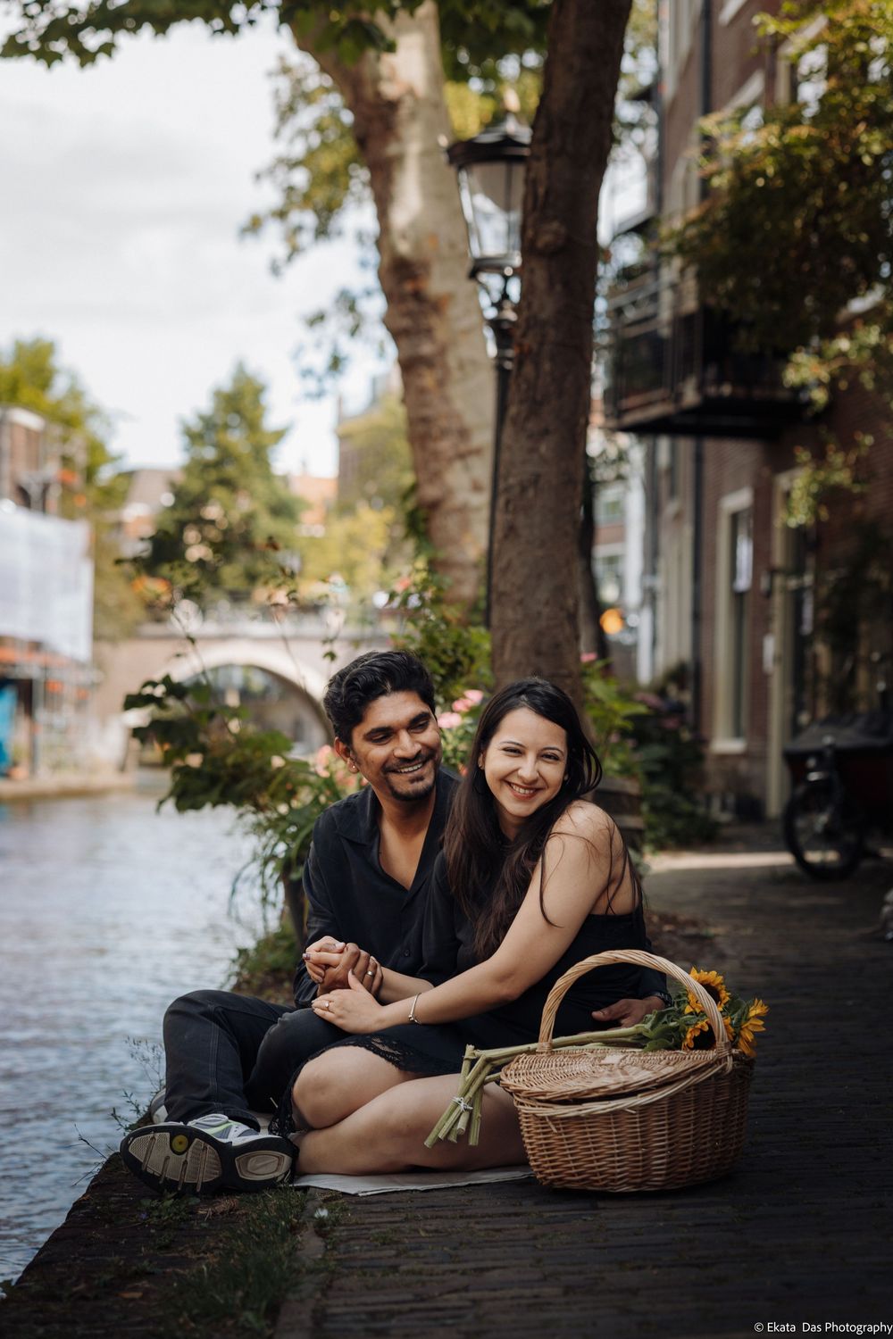 Couple sitting by a canal in Amsterdam, smiling and holding hands with a wicker basket of flowers nearby.