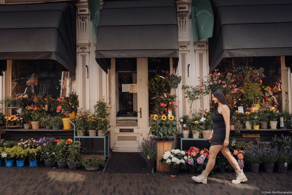 Woman in dark dress walks past a charming flower shop with colorful blooms displayed outside on a cobblestone street.