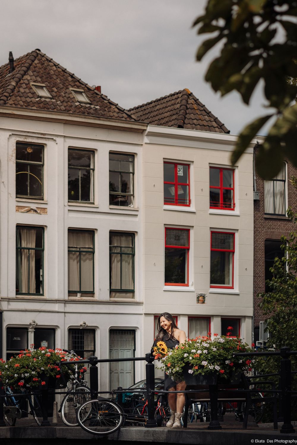 Woman posing on a bridge with sunflowers beside classic white Dutch canal houses with red-trimmed windows and bicycles in Amsterdam.
