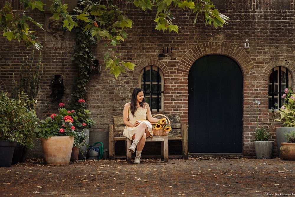 Woman in cream outfit sitting on wooden bench outside rustic brick building with arched door and potted plants.