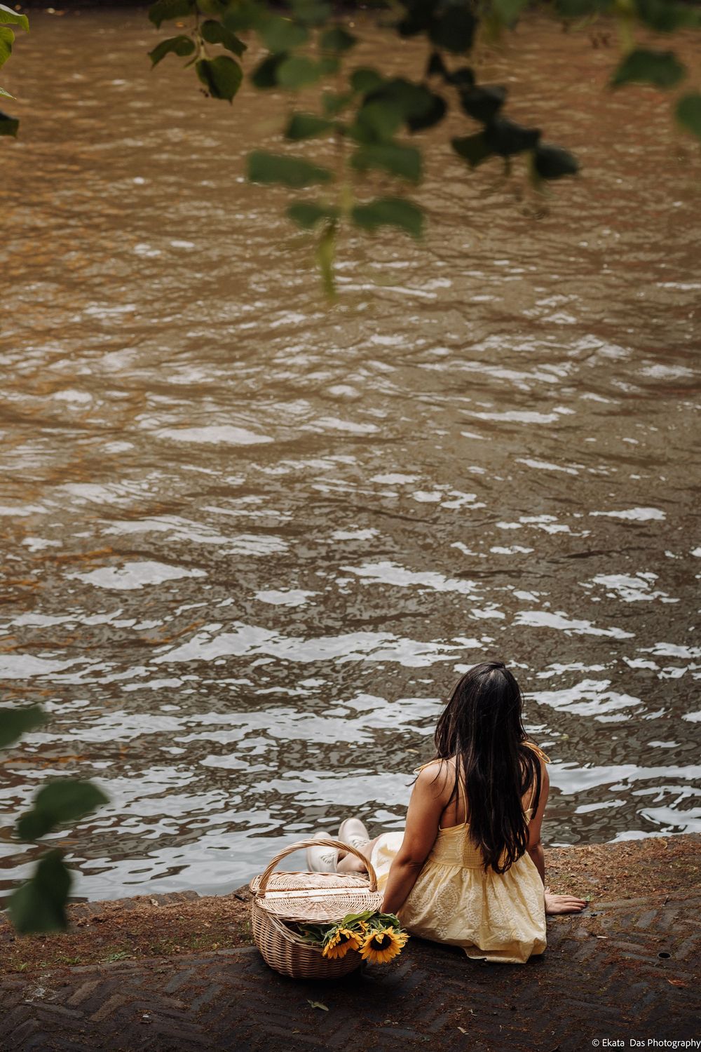 Young girl in yellow dress sits by a river with a wicker basket of sunflowers, viewed from above.