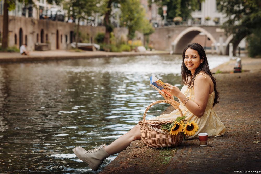 A smiling woman in a yellow dress sits by a canal with a picnic basket of sunflowers and a baguette.