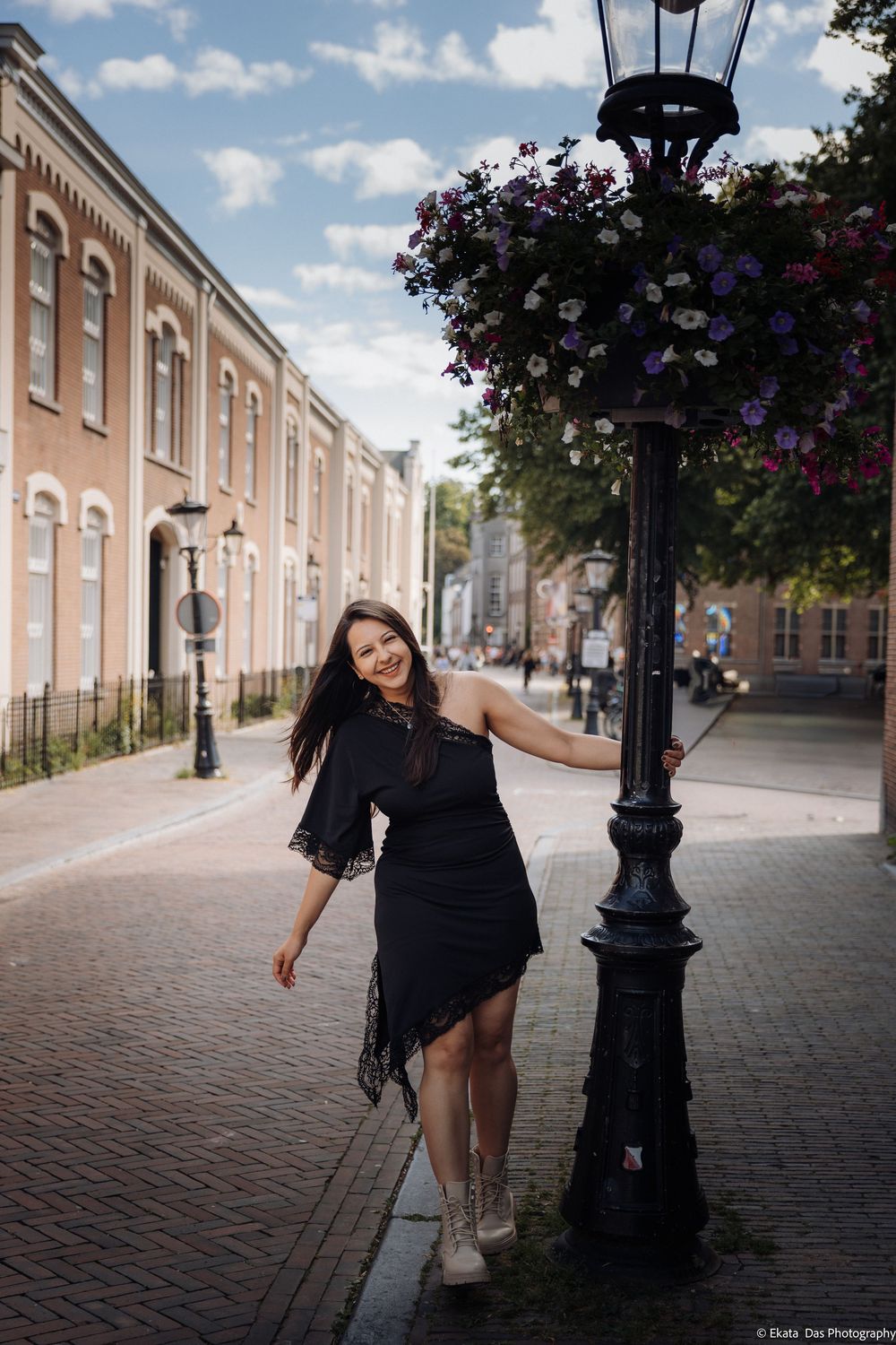 Woman in black dress posing by ornate lamppost with flowers on cobblestone street in historic European town.