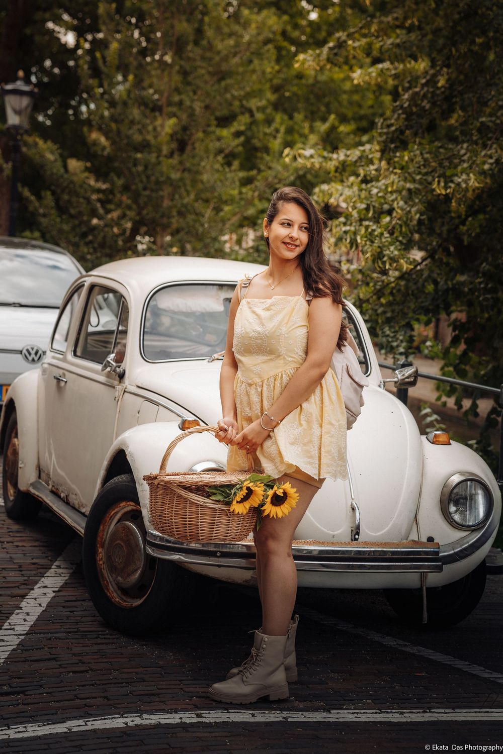 Young woman in yellow dress holding sunflowers basket, leaning against vintage white VW Beetle car outdoors.