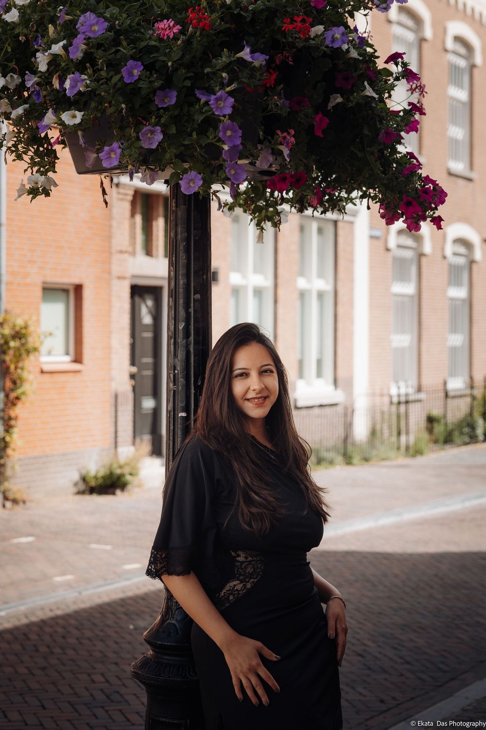 Woman in black dress smiling under hanging flower basket on a charming European brick street.